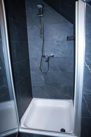 A shower with a handheld showerhead and a wall-mounted faucet in a room with dark tiled walls. - Angelis Pension Frankfurt Airport (Kelsterbach)