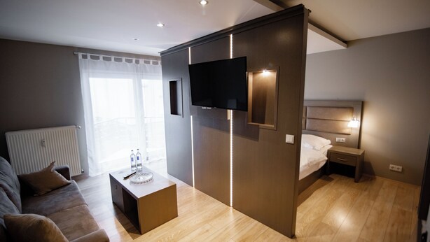A modern hotel room with a flat-screen TV, a bed, a bedside table, a sofa, and a wooden floor.