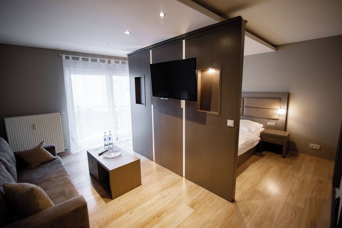 A modern hotel room with a flat-screen TV, a bed, a bedside table, a sofa, and a wooden floor.
