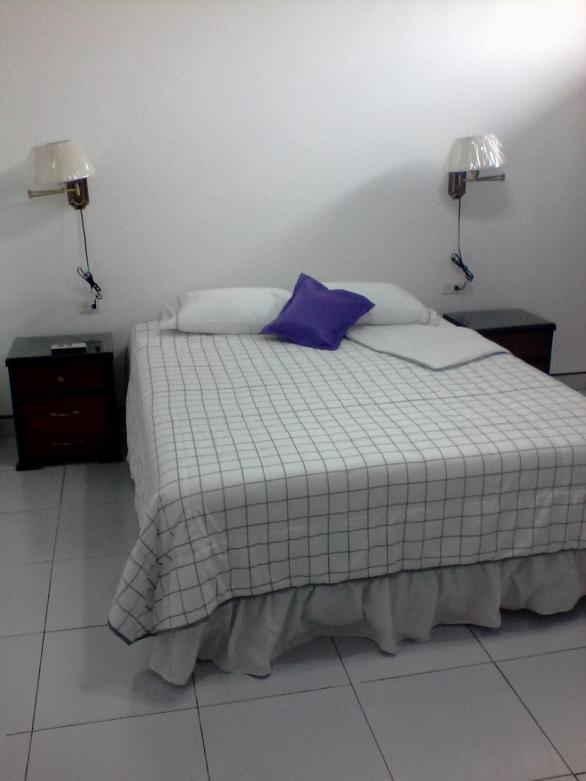 A neatly made bed with a checkered bedspread and a single purple pillow, flanked by two bedside tables with lamps.