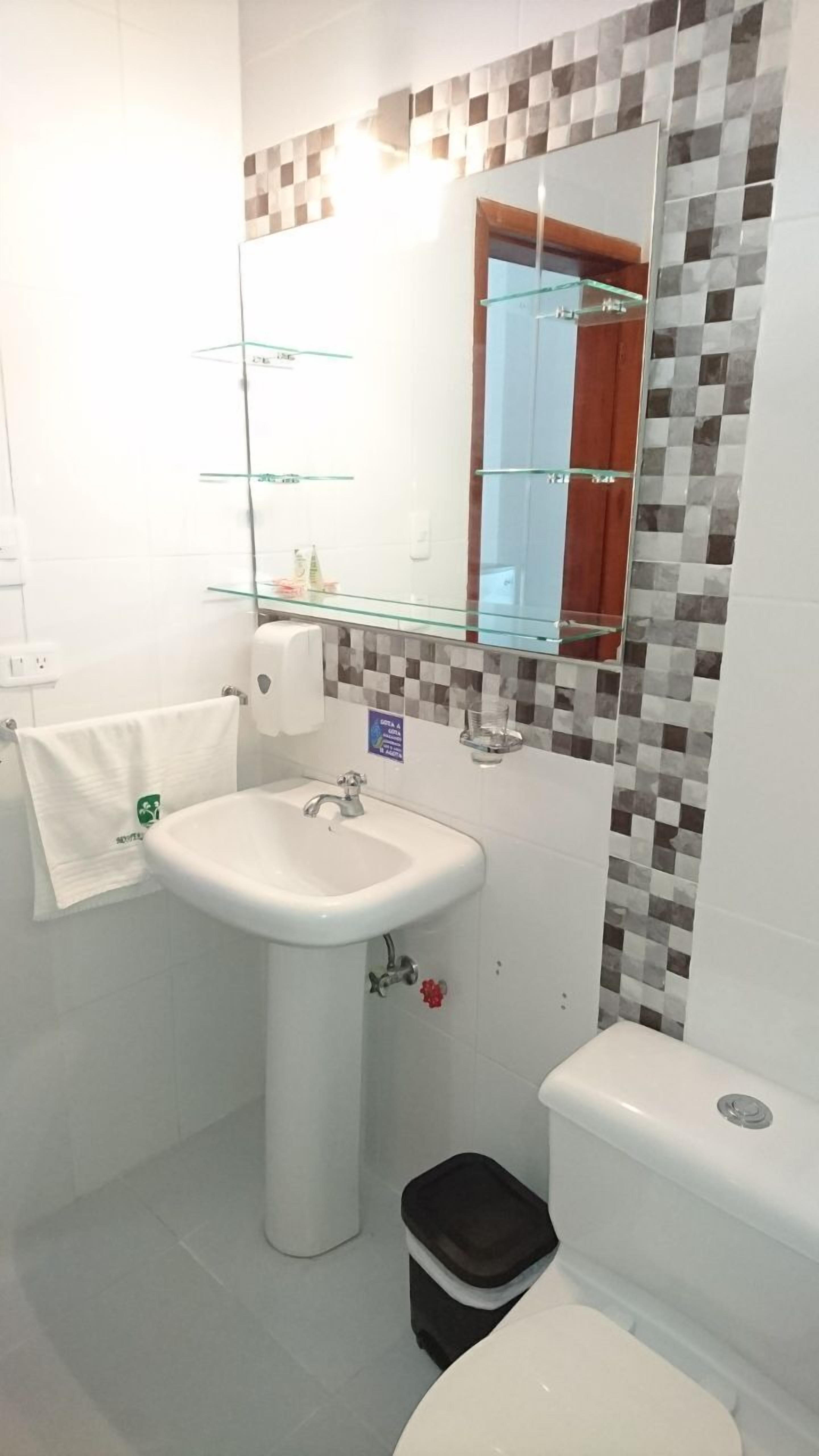 A bathroom with a white sink, a mirror, and a toilet. There is a glass shelf above the sink and a small trash bin next to the toilet.