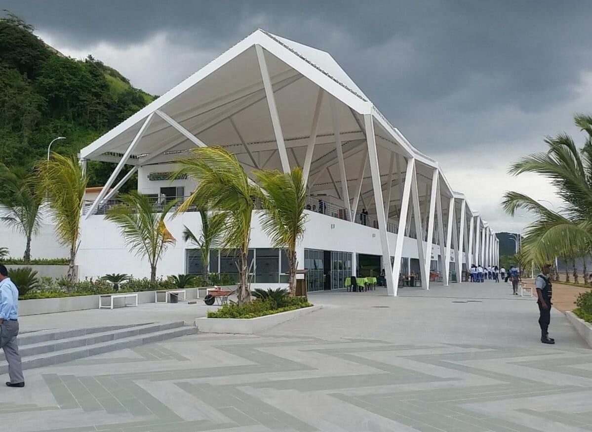 A modern building with a white, geometric roof structure and a paved plaza with palm trees.