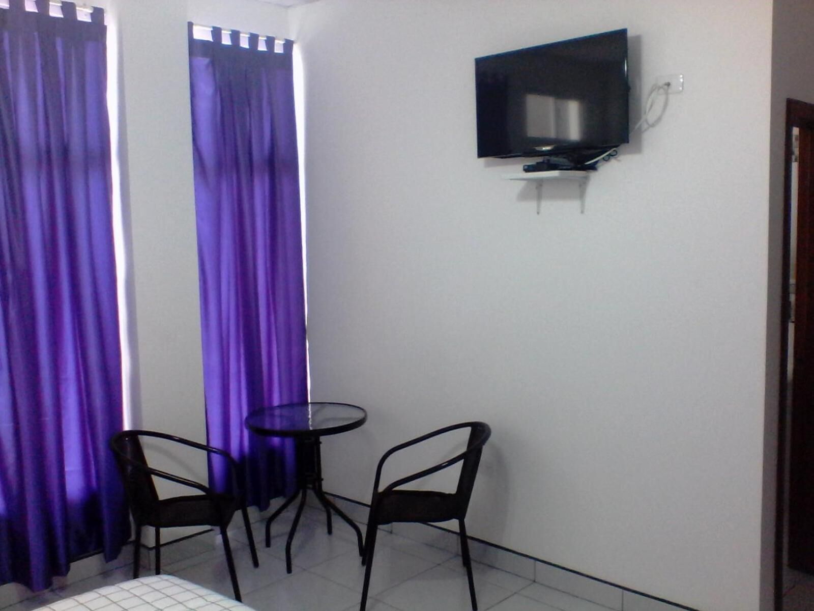 Une petite pièce avec un téléviseur fixé au mur, deux chaises, une petite table ronde et des rideaux violets.