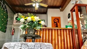 Una habitación con ventilador de techo, una mesa de madera con mantel de encaje, un florero con flores y un fondo de bambú.