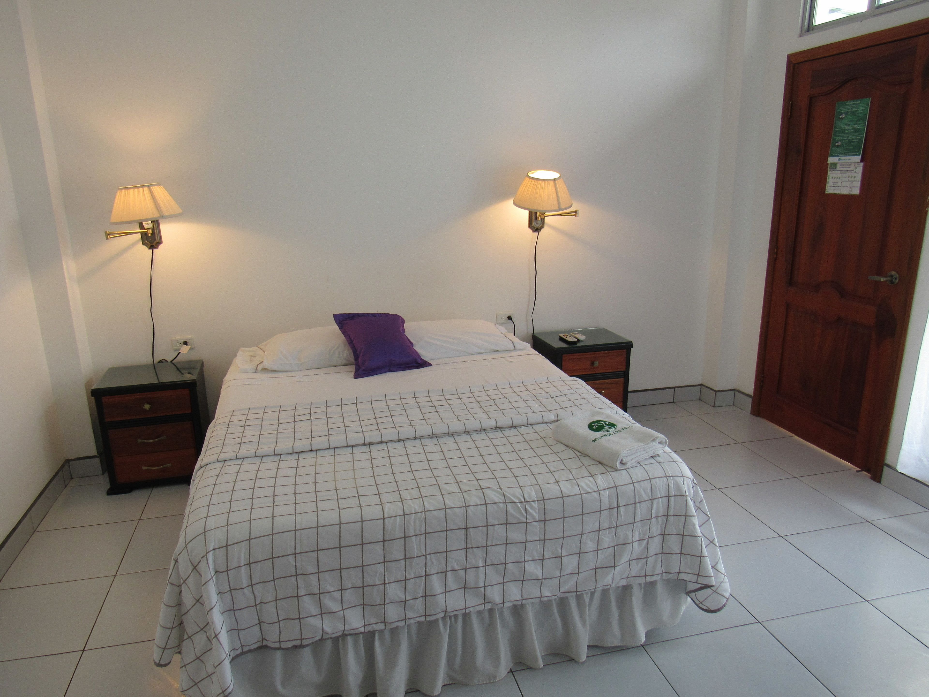 Una cama bien hecha con una colcha a cuadros, dos lámparas de noche, una almohada morada y una toalla doblada en el suelo.