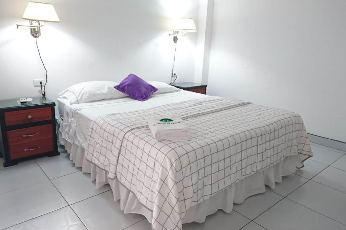 A neatly made bed with a checkered bedspread, a purple pillow, and a folded towel on the bed. There are two bedside tables with lamps, one on each side of the bed.