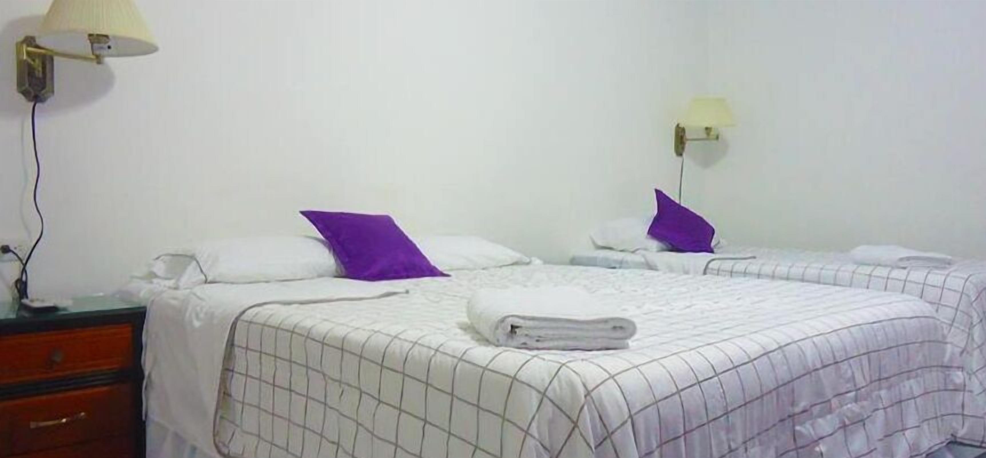 Habitación de hotel con dos camas, cada una con cubrecama blanco y almohadas moradas, una mesita de noche y una lámpara.