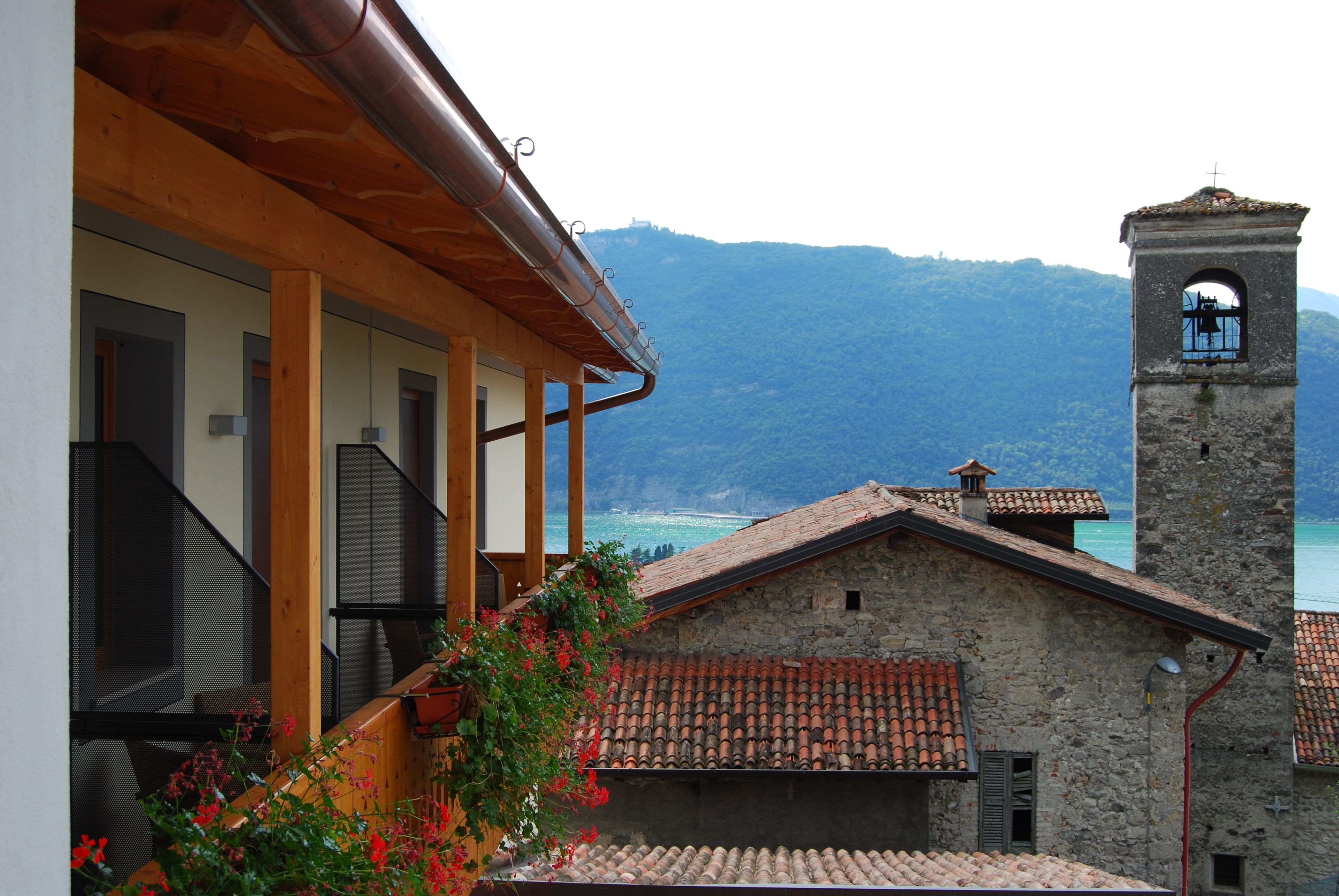 A stone building with a bell tower, wooden balcony, and a view of the sea and mountains.