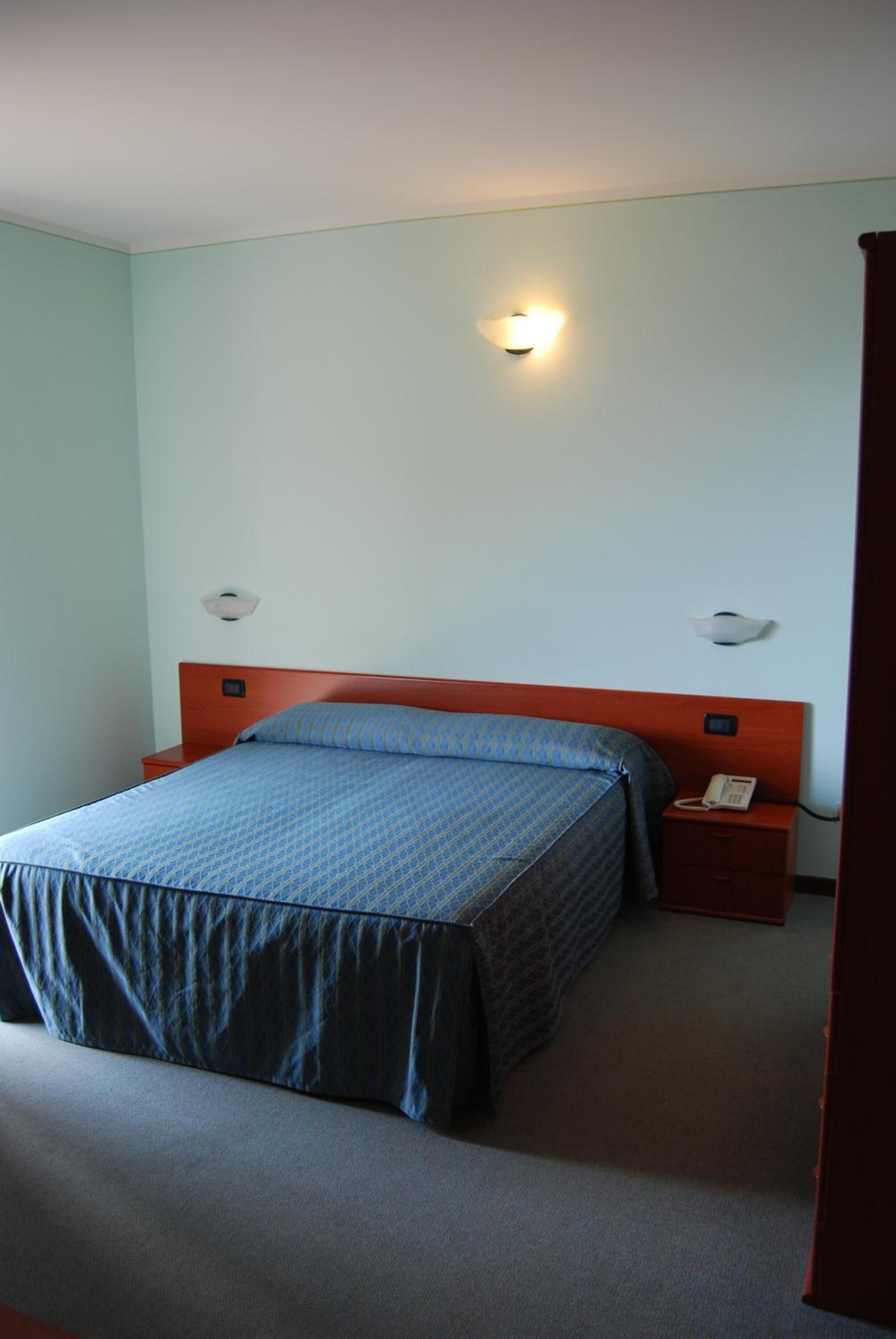 Una cama individual con cabecera de madera, una mesita de noche con teléfono, y una lámpara fijada a la pared.