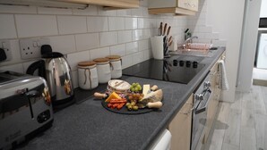 Une cuisine avec un plan de travail noir, des carreaux blancs, un grille-pain, une bouilloire, un porte-couteaux et une planche à fromage avec divers fromages et fruits.