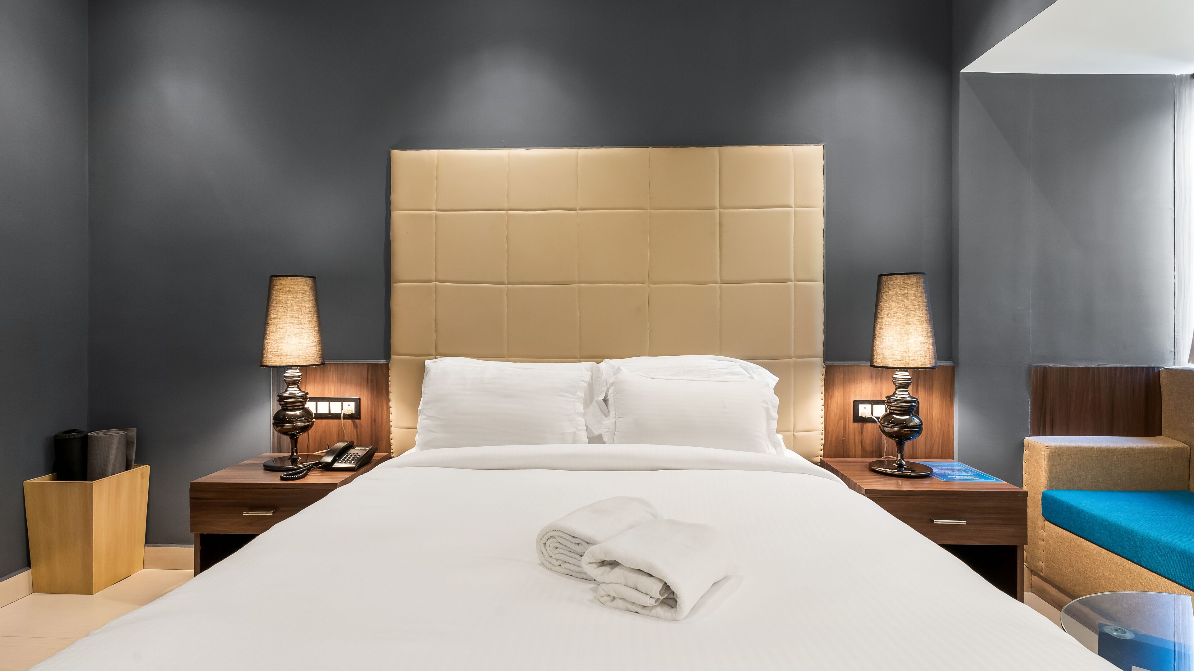 Habitación de hotel con una cama grande, dos mesitas de noche con lámparas, un cabecero de madera y una silla azul.