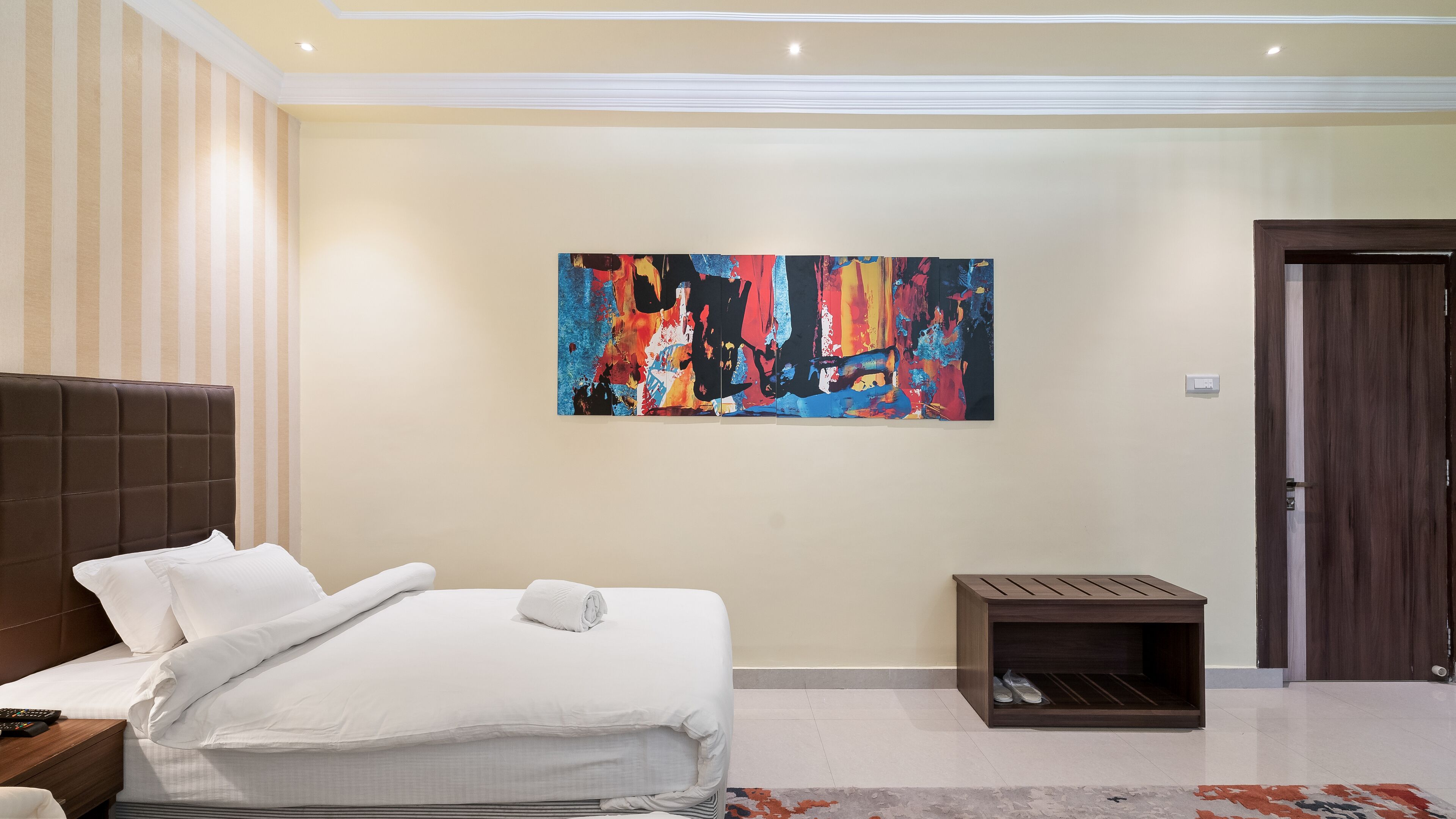 Un dormitorio moderno con una cama grande, una mesita de noche, un banco de madera y un cuadro abstracto de colores en la pared.
