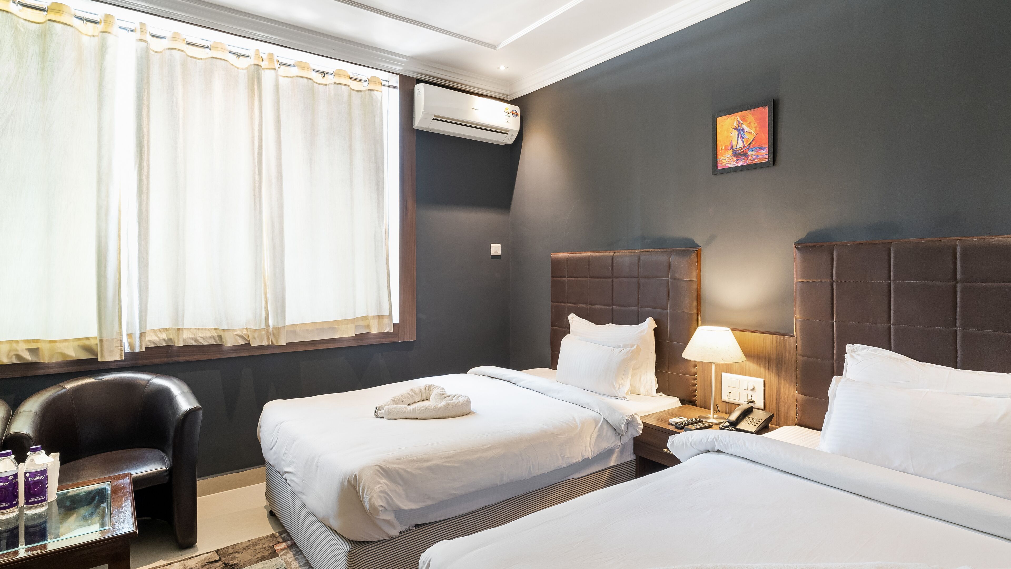 A hotel room with two beds, a chair, a table with bottles, an air conditioner, and a painting on the wall.