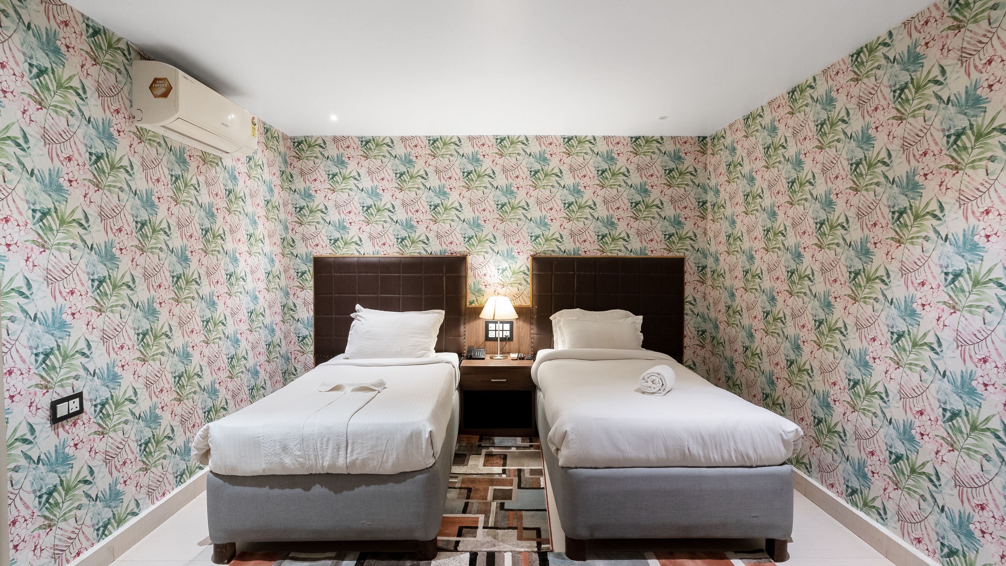 A room with two beds, a nightstand with a lamp, and floral wallpaper.