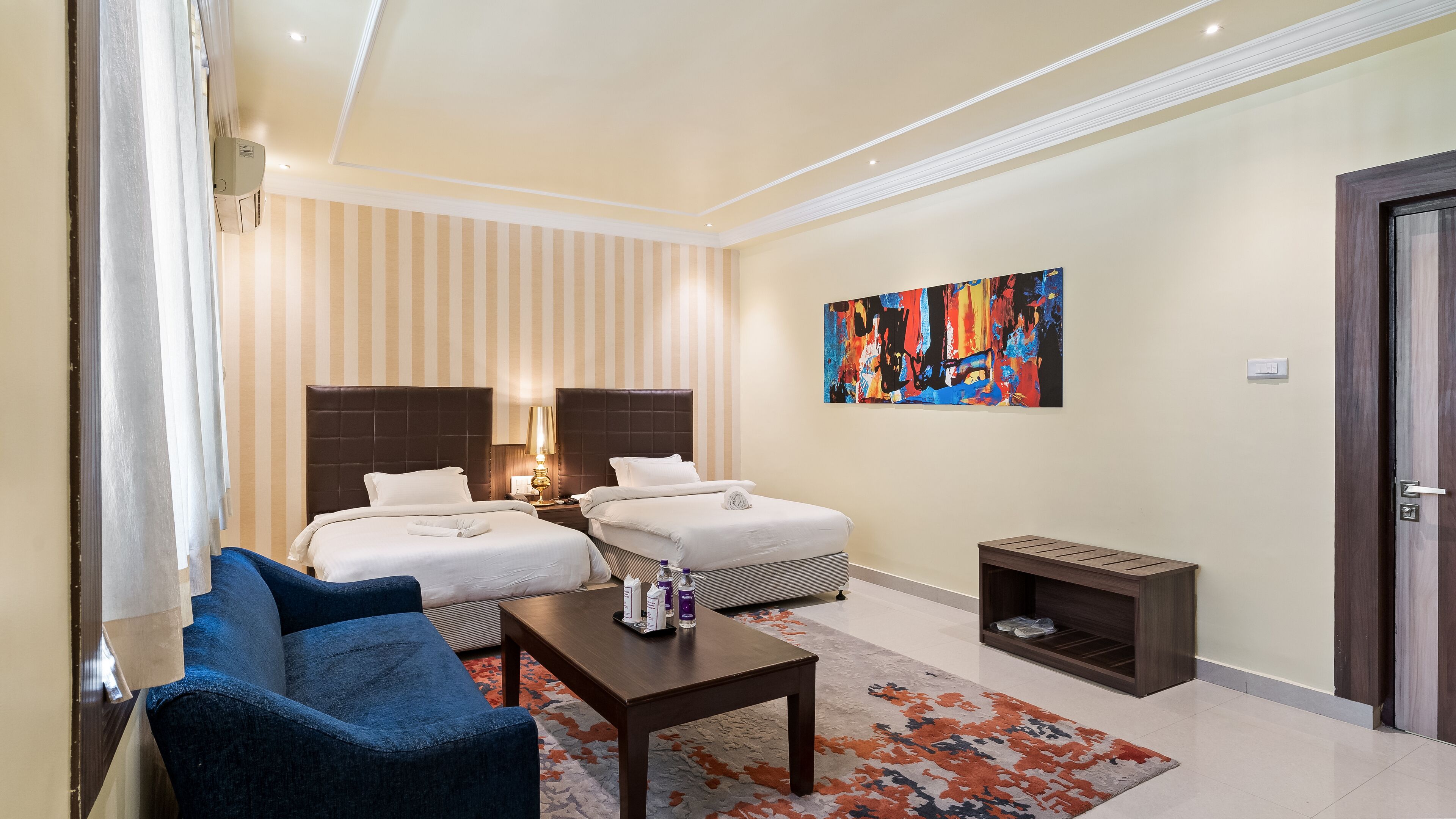 A hotel room with two beds, a small table, a chair, a colorful abstract painting, and a shoe shelf.
