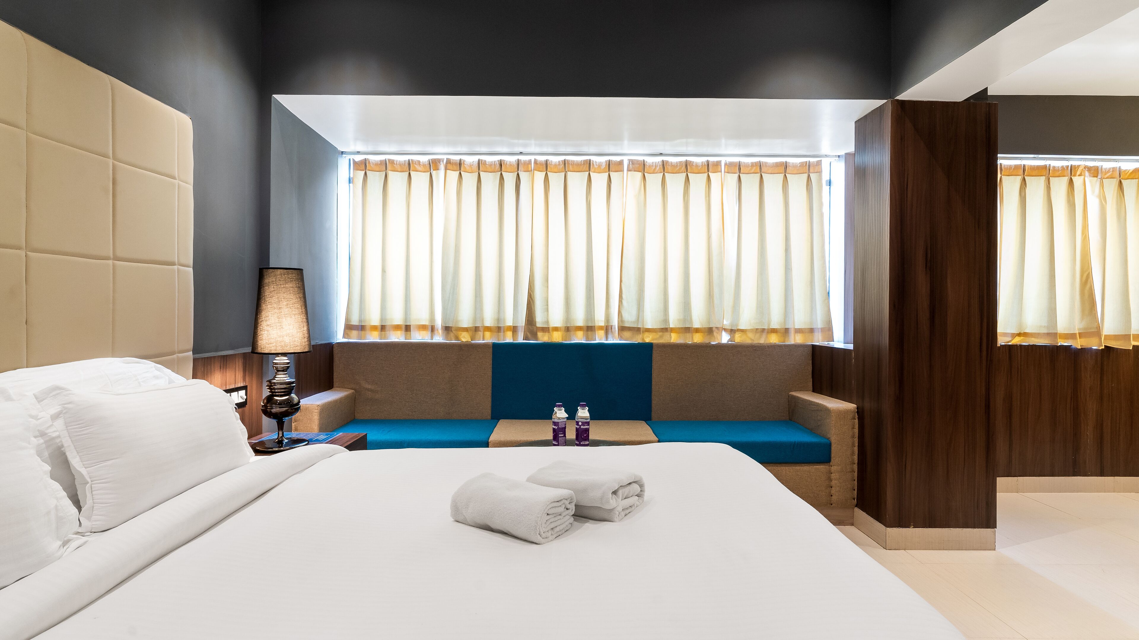 Una habitación de hotel moderna con una cama grande, una mesita de noche con lámpara, una ventana con cortinas y un banco tapizado en azul.