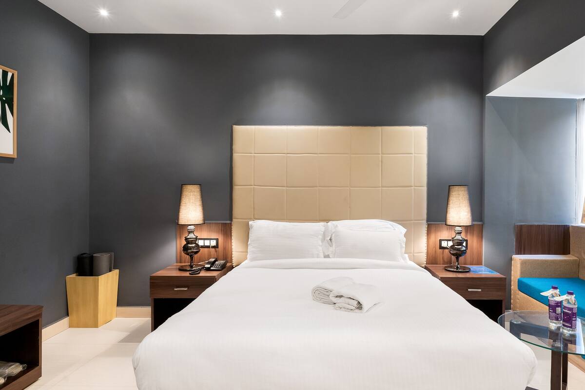 Een moderne hotelkamer met een groot bed, twee nachtkastjes met lampen, een glazen salontafel, een blauwe bank en uitzicht op het omliggende landschap.