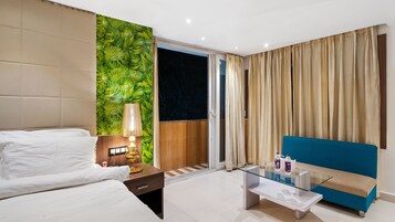 Habitación de hotel con cama, mesita de noche con lámpara, mesa de vidrio con artículos de aseo, sofá azul y pared cubierta de hojas verdes.