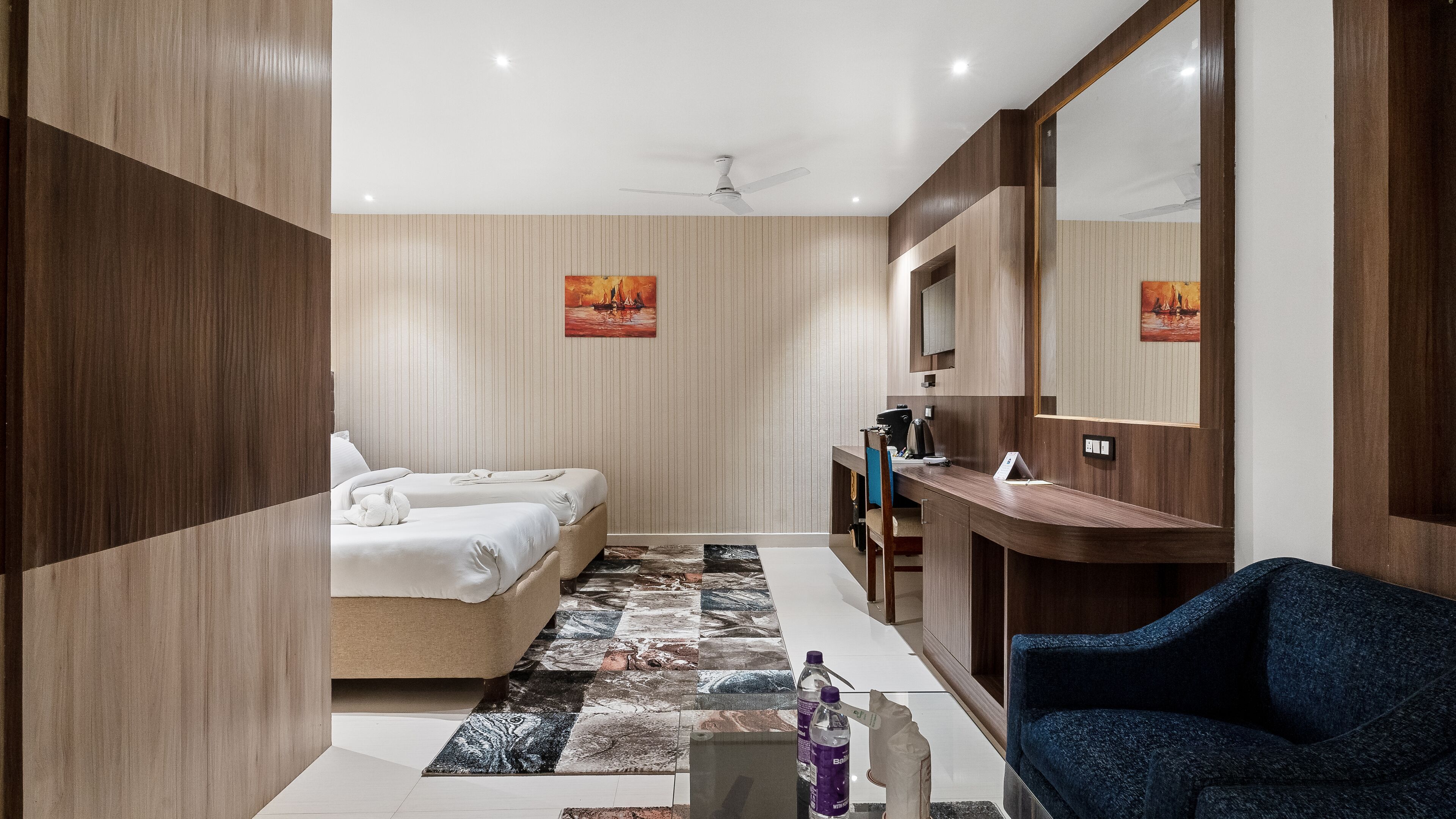 A hotel room with two beds, a desk, a chair, a mirror, and a ceiling fan.