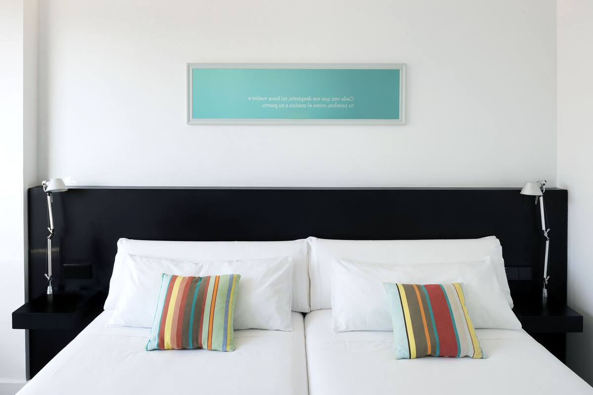 A double bed with white bedding and colorful striped pillows, a black headboard, and a teal framed quote on the wall.