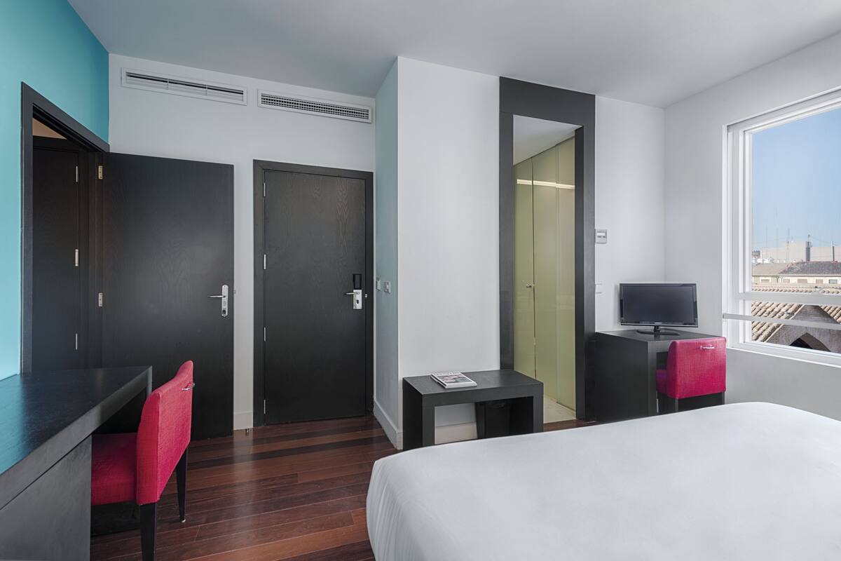 A hotel room with a bed, a desk with a chair, a television, and a window with a city view.