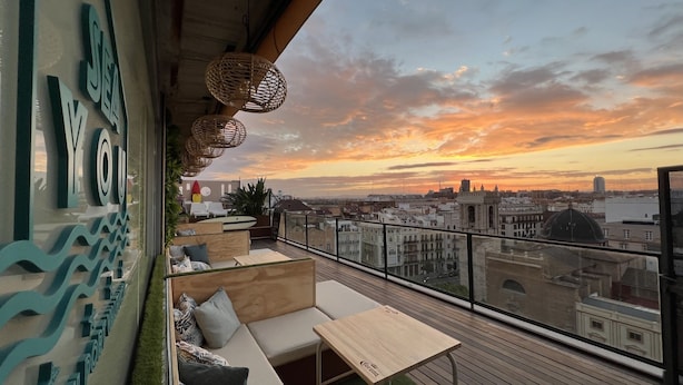 Terraza en la azotea con vistas a la ciudad al atardecer, que cuenta con un piso de madera, zona de estar y lámparas de mimbre decorativas.