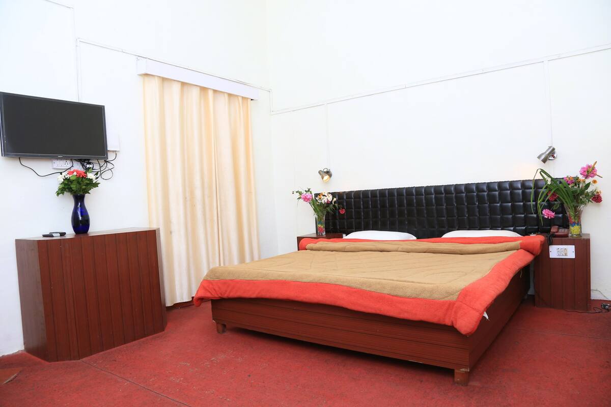 A hotel room with a bed, a TV mounted on the wall, a bedside table with a vase of flowers, and a window with curtains.