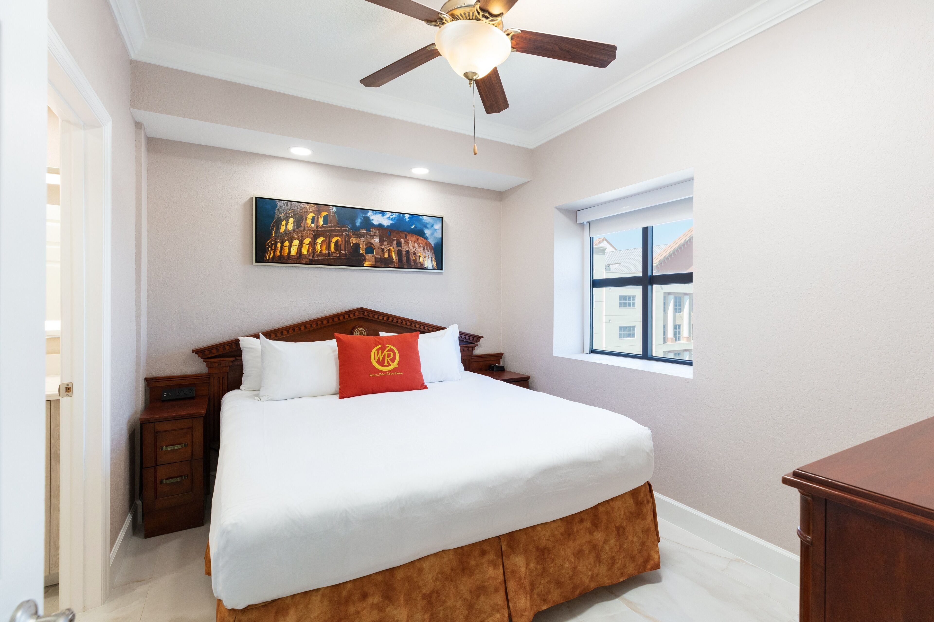 A bedroom with a large bed, a ceiling fan, a window with a view of buildings, and a painting of a historical building on the wall.