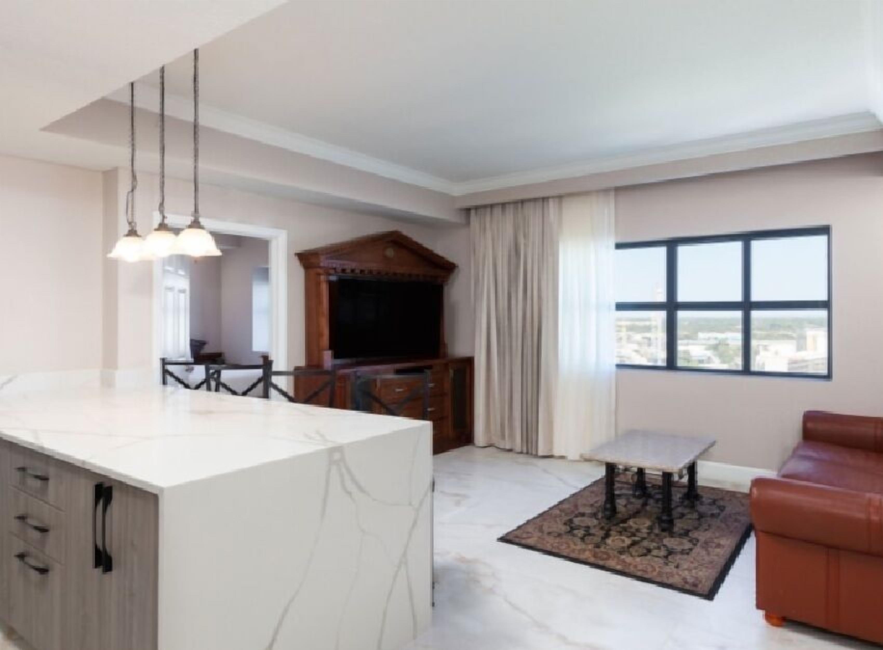 A modern living room with a marble island, a dining area, a brown leather sofa, and a large window with a city view.