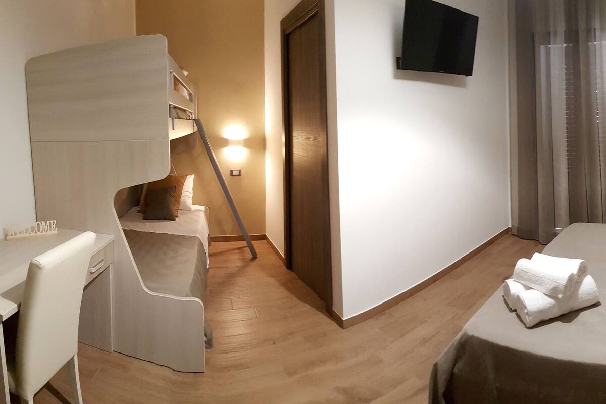 Une chambre d’hôtel avec un lit superposé, un bureau avec une chaise, un lit avec des oreillers, un téléviseur à écran plat et un tableau au mur.