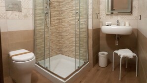 A bathroom with a glass-enclosed shower, a toilet, a sink, and a small stool. - B&B Il Campanile (Sciacca)