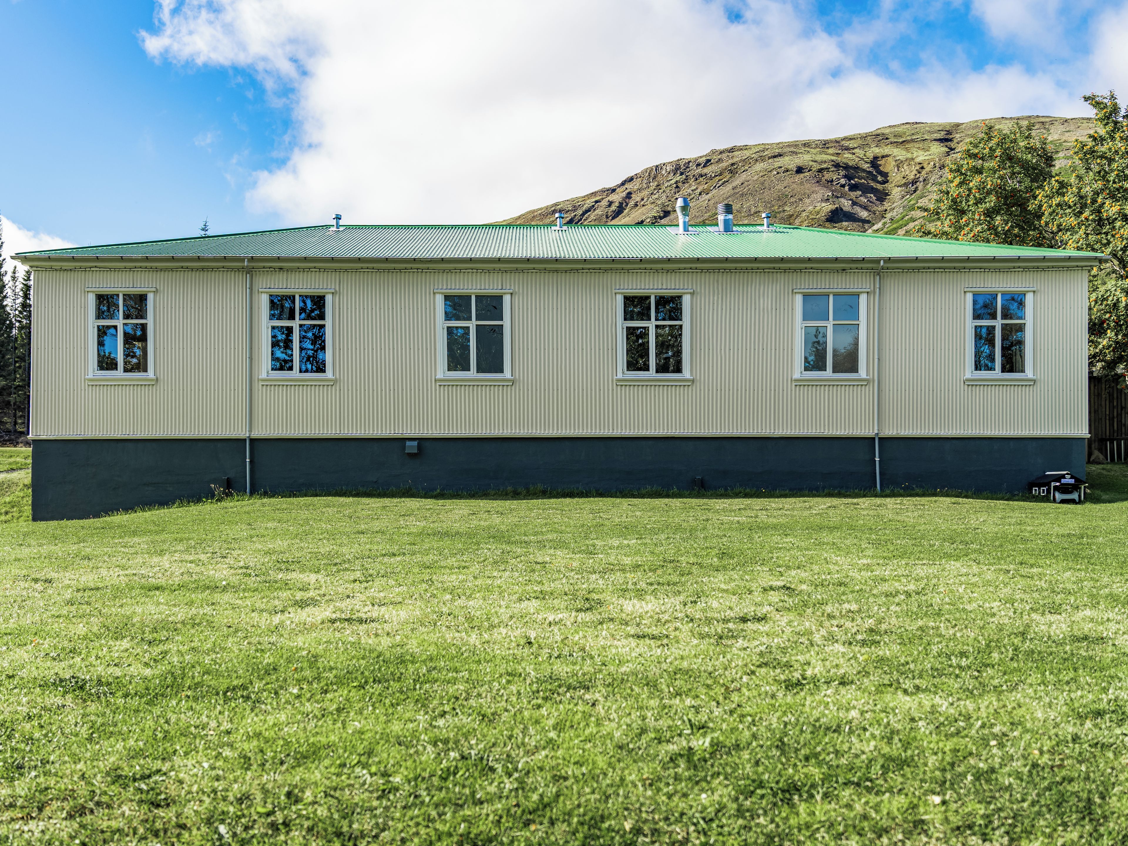 A two-story building with a green roof and white siding, situated on a grassy field with a hill in the background.