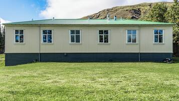 A two-story building with a green roof and white siding, situated on a grassy field with a hill in the background.