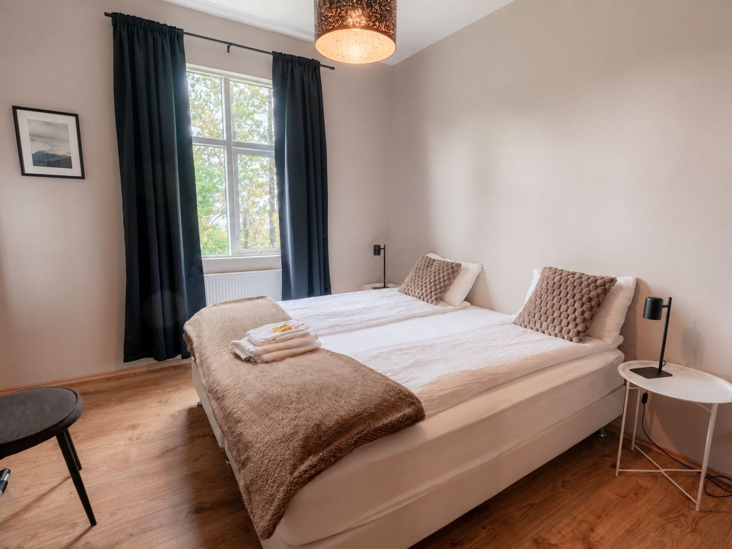 Un dormitorio con una cama grande, una mesita de noche, una silla, una ventana con cortinas y una lámpara colgante.
