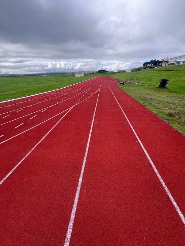 Pista de atletismo roja con marcas blancas, rodeada de campos verdes y un cielo nublado.