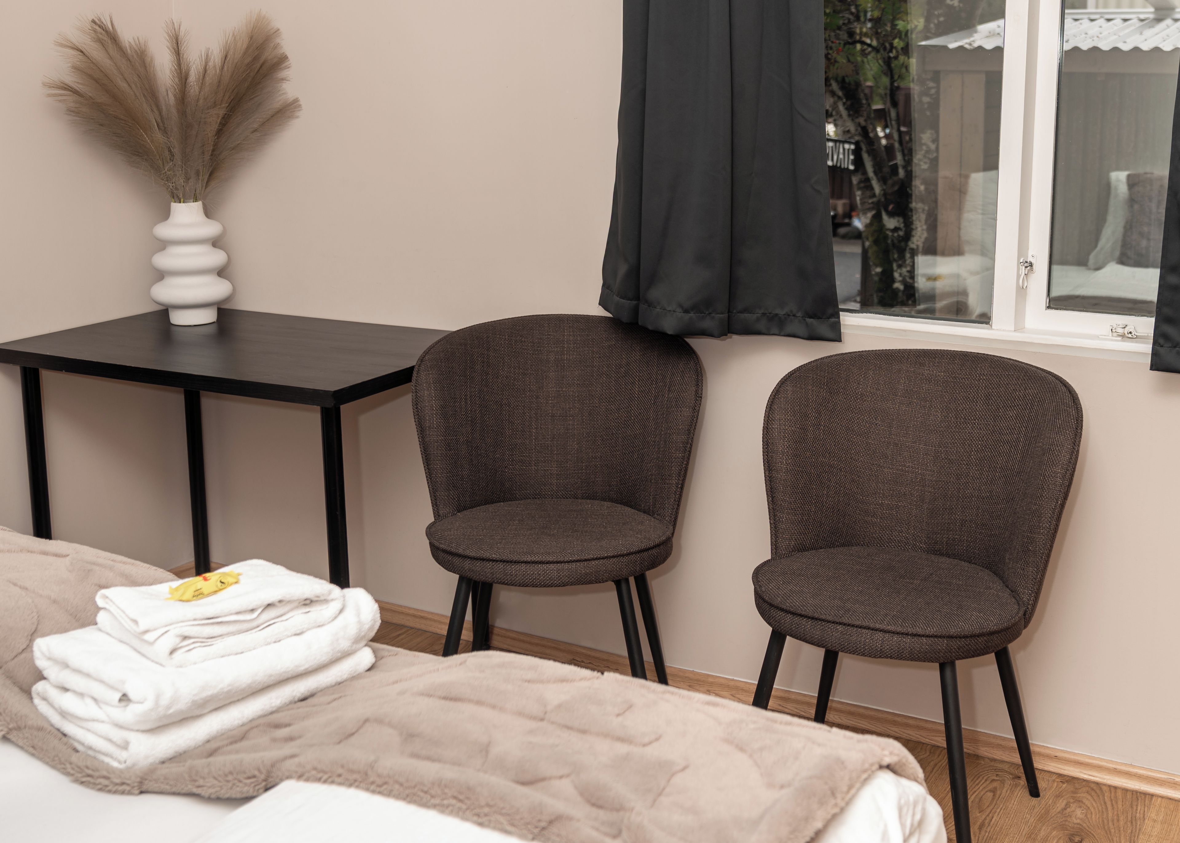 A room with a bed, two chairs, a table, a vase with dried plants, and a window with curtains.