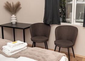 Una habitación con cama, dos sillas, una mesa, un jarrón con plantas secas y una ventana con cortinas.
