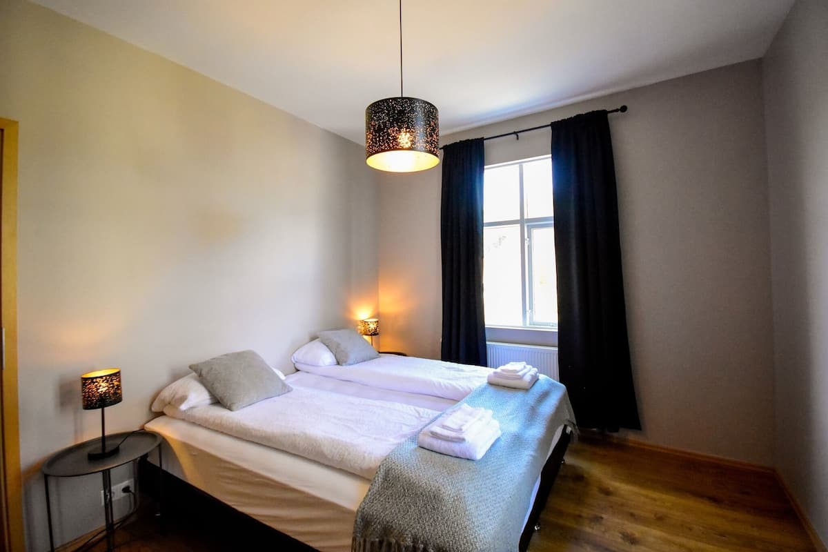 Una cama doble con una manta gris, una lámpara de noche y una ventana con cortinas oscuras.