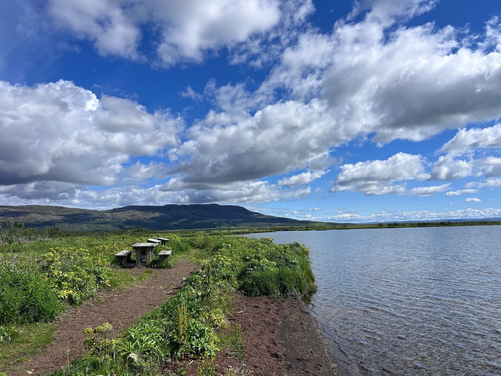 A lakeside view with a clear sky, a path leading to a small structure, and distant mountains.