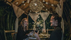 A couple dining at an outdoor table with a rustic wooden ceiling and hanging lights, surrounded by greenery.