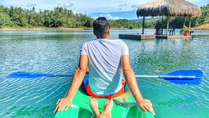 A man paddling a green kayak on a calm lake with a thatched-roof structure in the background.