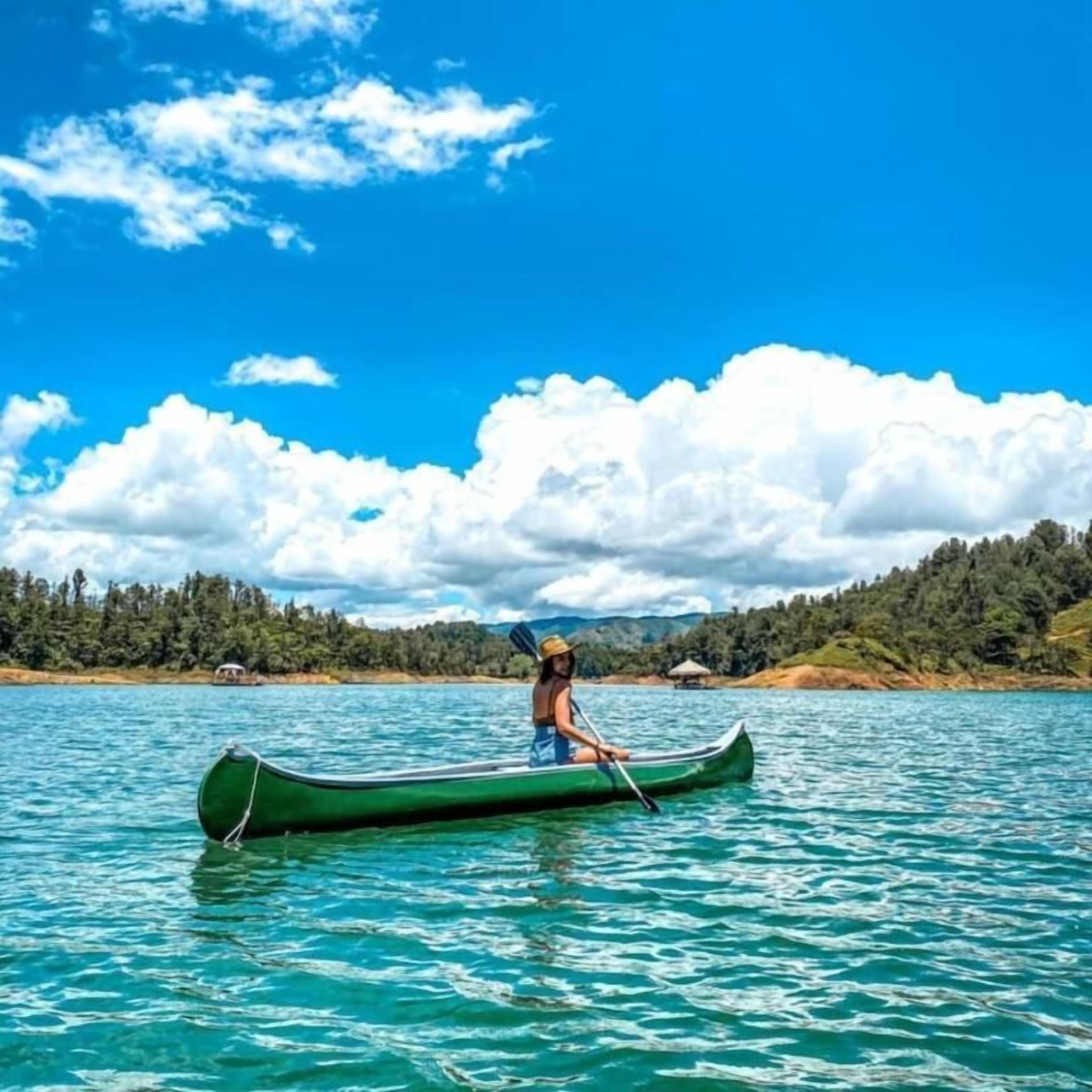 A person in a green canoe on a calm body of water with a forested shoreline and a clear sky.