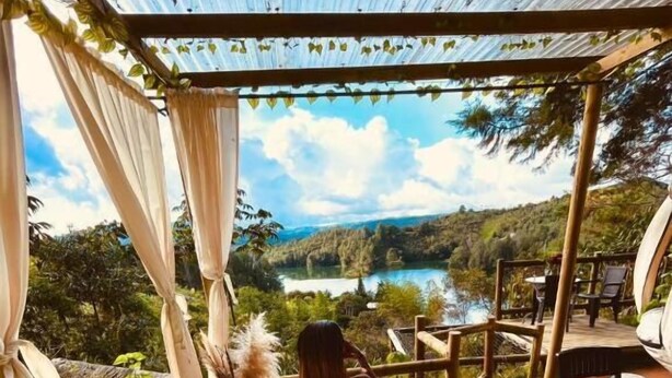 A hot tub with a person sitting inside, surrounded by a wooden deck and a scenic view of a lake and hills.