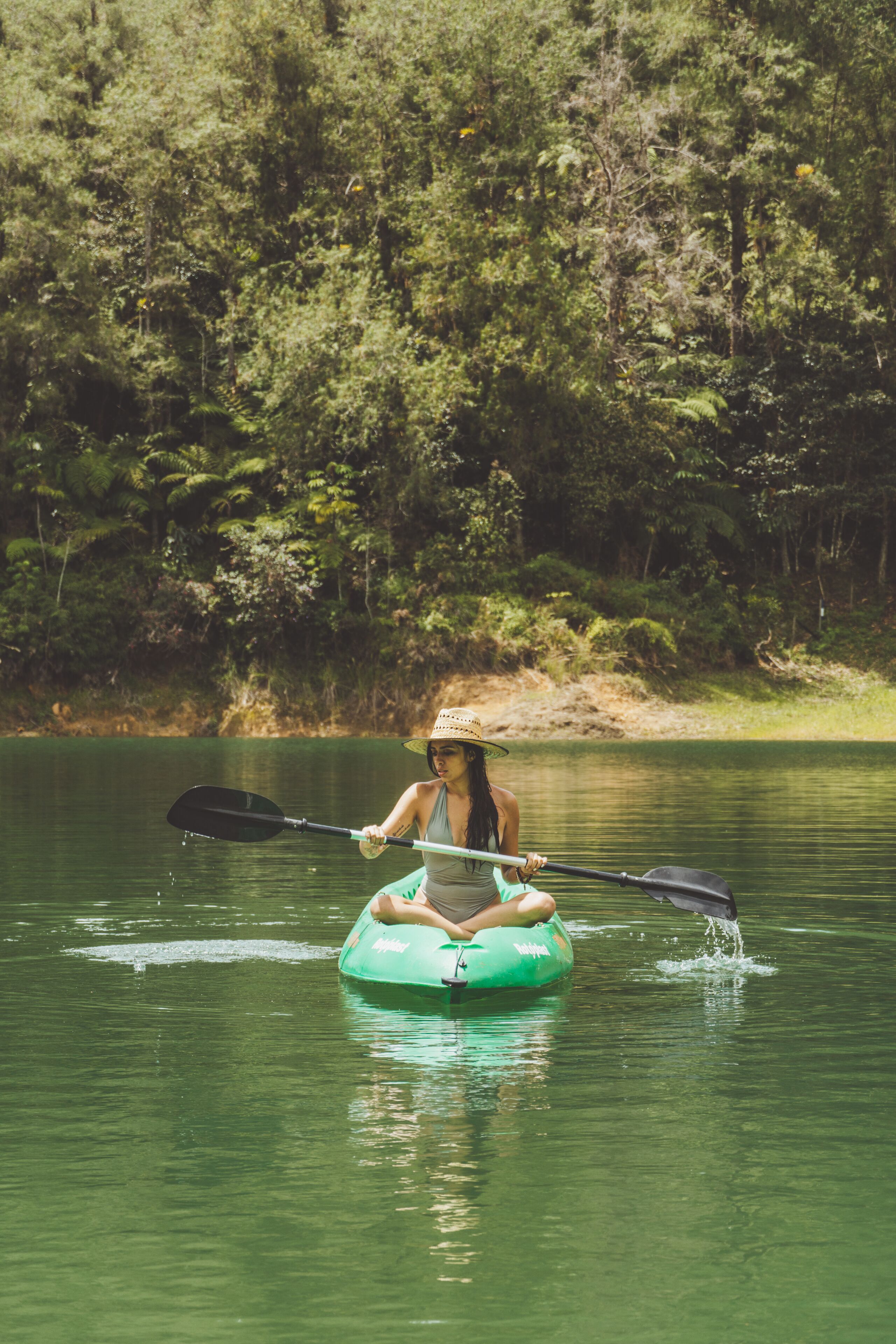 A person kayaking on a green inflatable raft in a calm body of water surrounded by dense greenery.