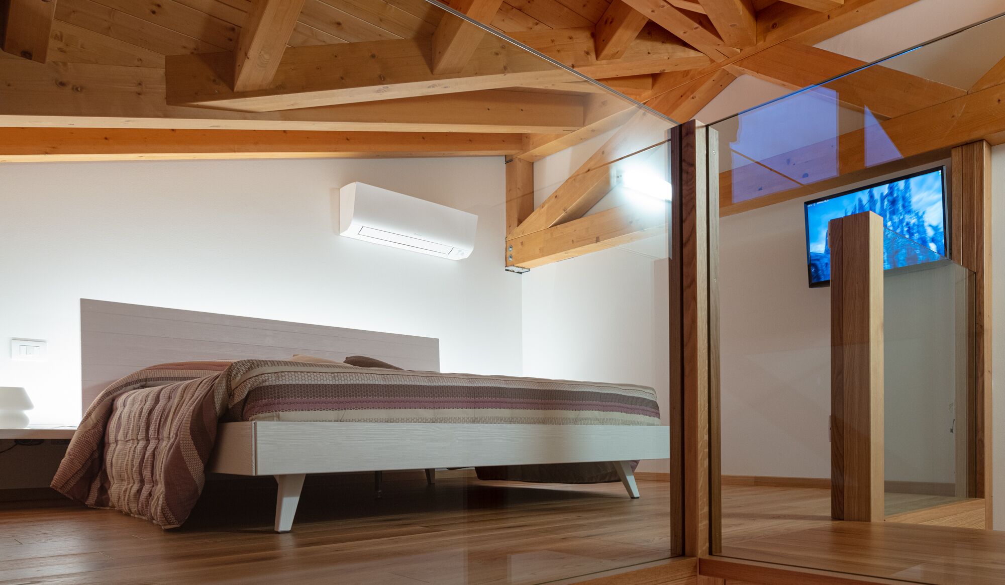 Ein modernes Schlafzimmer mit Holzbalkendecke, einem Bett mit einem gemusterten Überwurf, einer Wand-Klimaanlage und einer Glastür, die auf einen Balkon mit Blick führt.