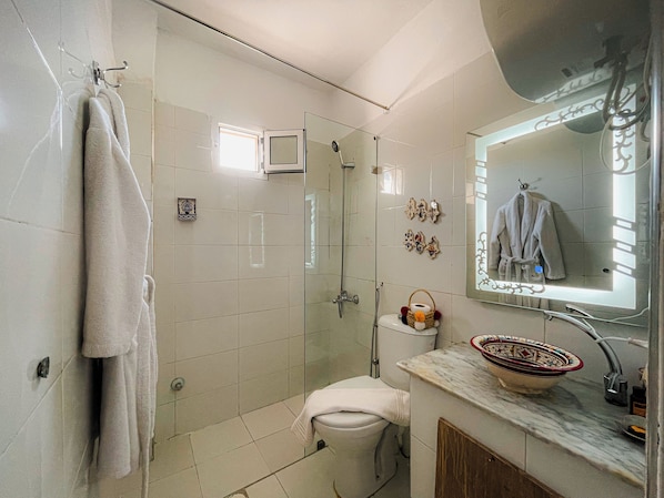 Ein Badezimmer mit Duschkabine aus Glas, Toilette, Waschbecken mit dekorativer Schüssel und Spiegel mit aufgehängtem Handtuch.