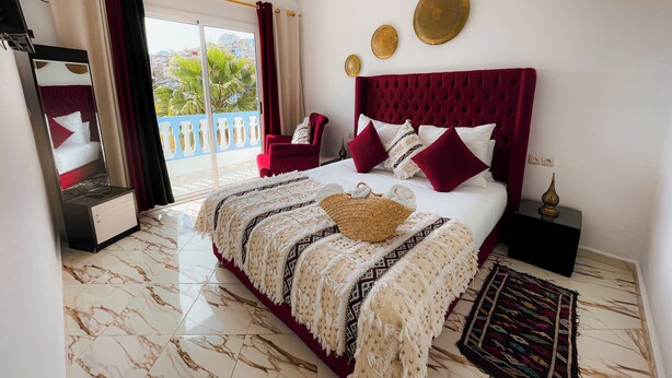 Quarto de hotel com cama, uma cadeira vermelha, uma janela grande e varanda.