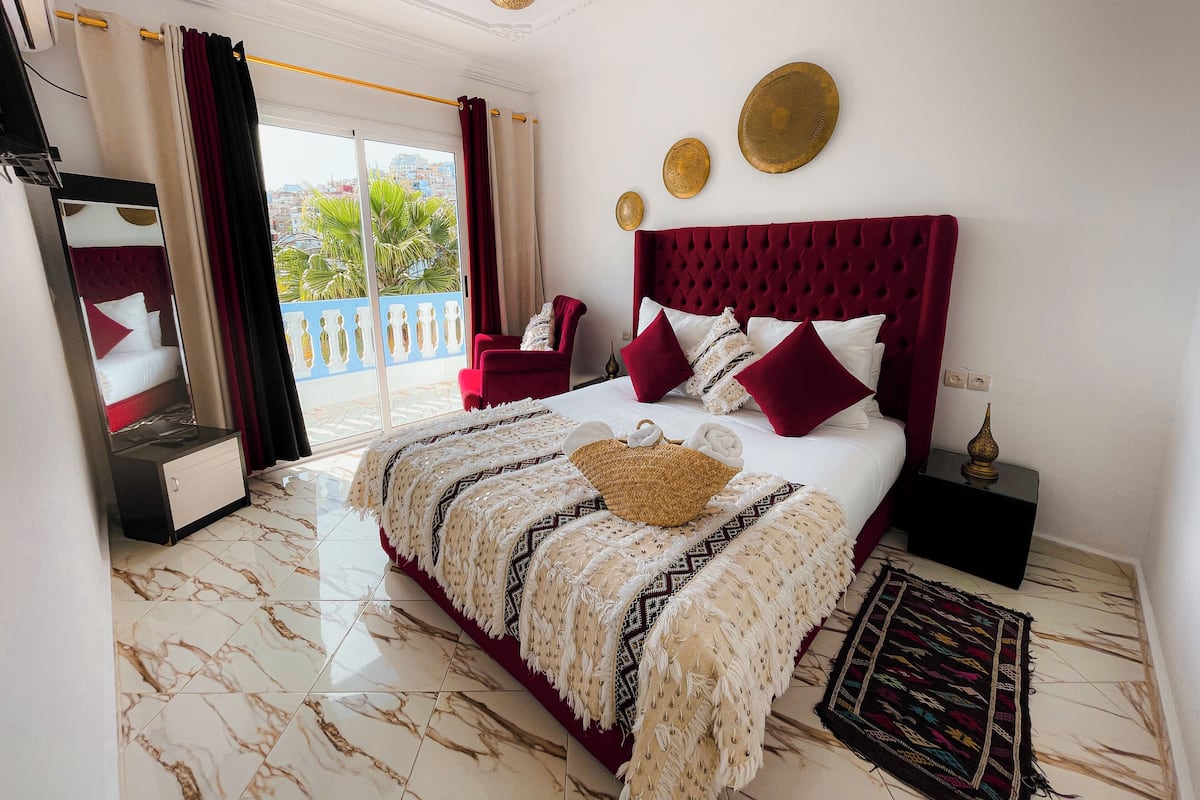 A hotel room with a bed, a red chair, a large window, and a balcony.