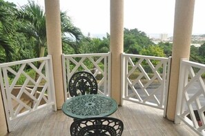 Un balcón con una mesa y sillas, rodeado de barandillas de celosía blanca, con vistas a un paisaje verde y exuberante.