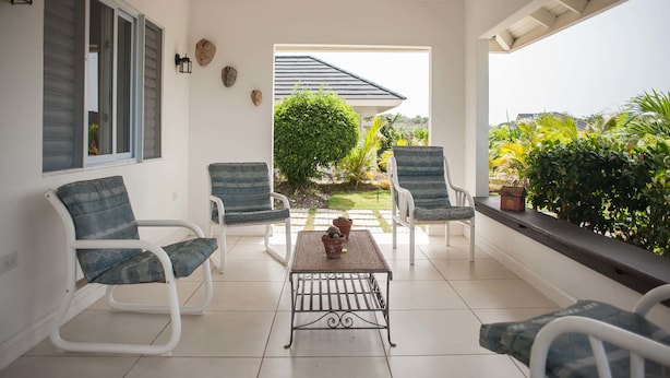 Un patio cubierto con sillas y una mesa blancos, una ventana con contraventanas y vistas a un jardín.